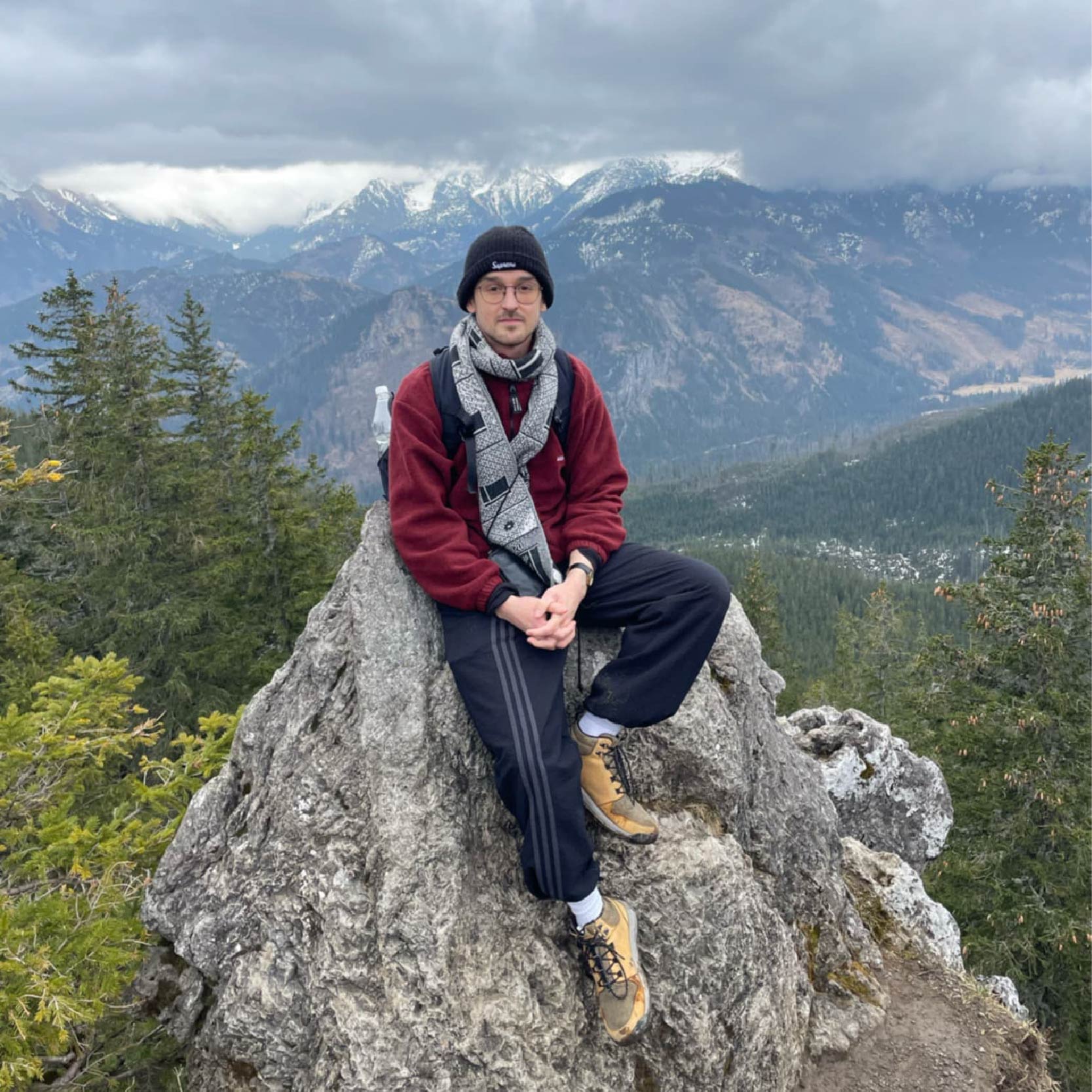 Photo of Jakub Cacek, PhD student, sitting on a rock outdoors, with mountains and greenery in the background. He is wearing a burgundy jacket, grey scarf, navy trousers, and a black beanie.