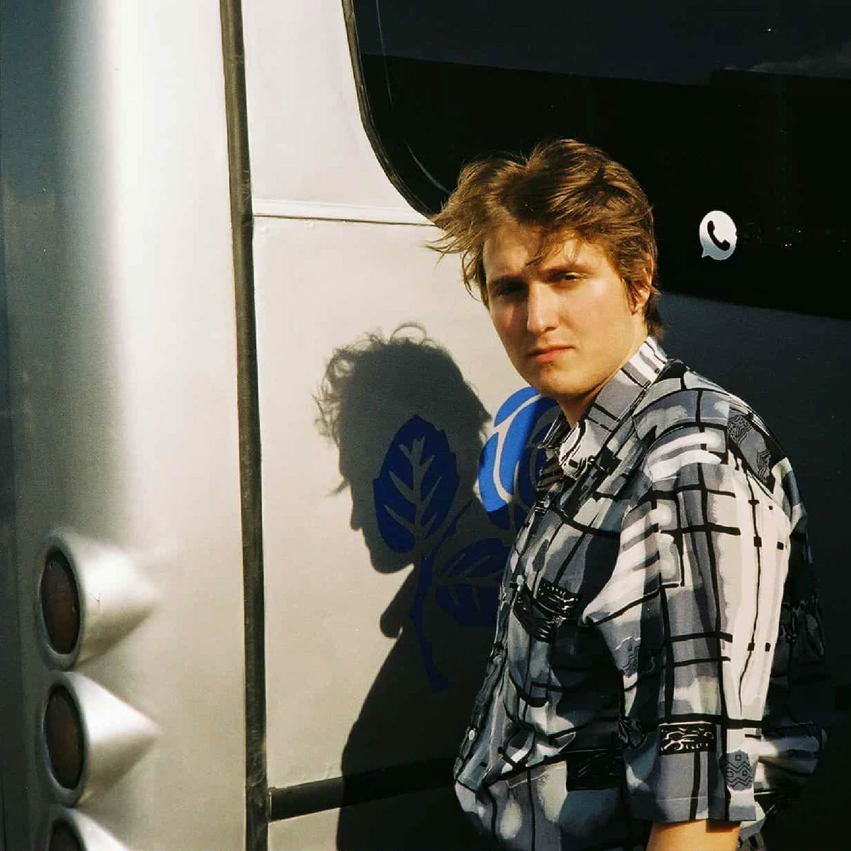 Photo of Wiktor Zieliński, PhD student, with dark blond hair, wearing a checked shirt and standing in sunlight, with a grey coach bus in the background.
